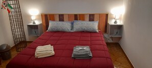 A bed with a red quilt, two pillows, two bedside lamps, a wooden headboard, and a small trash bin.