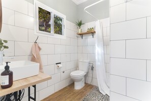 A bathroom with white tiles, a wooden vanity, a toilet, a shower curtain, and a window with a view of trees.