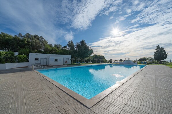 A rectangular swimming pool with a tiled deck, surrounded by greenery and a clear sky.
