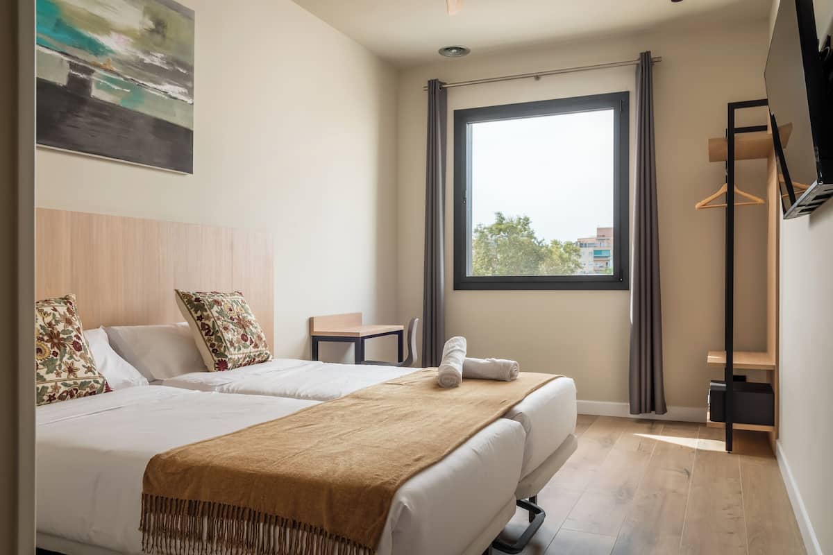 A hotel room with two beds, a large window, a small table, and a TV mounted on the wall.