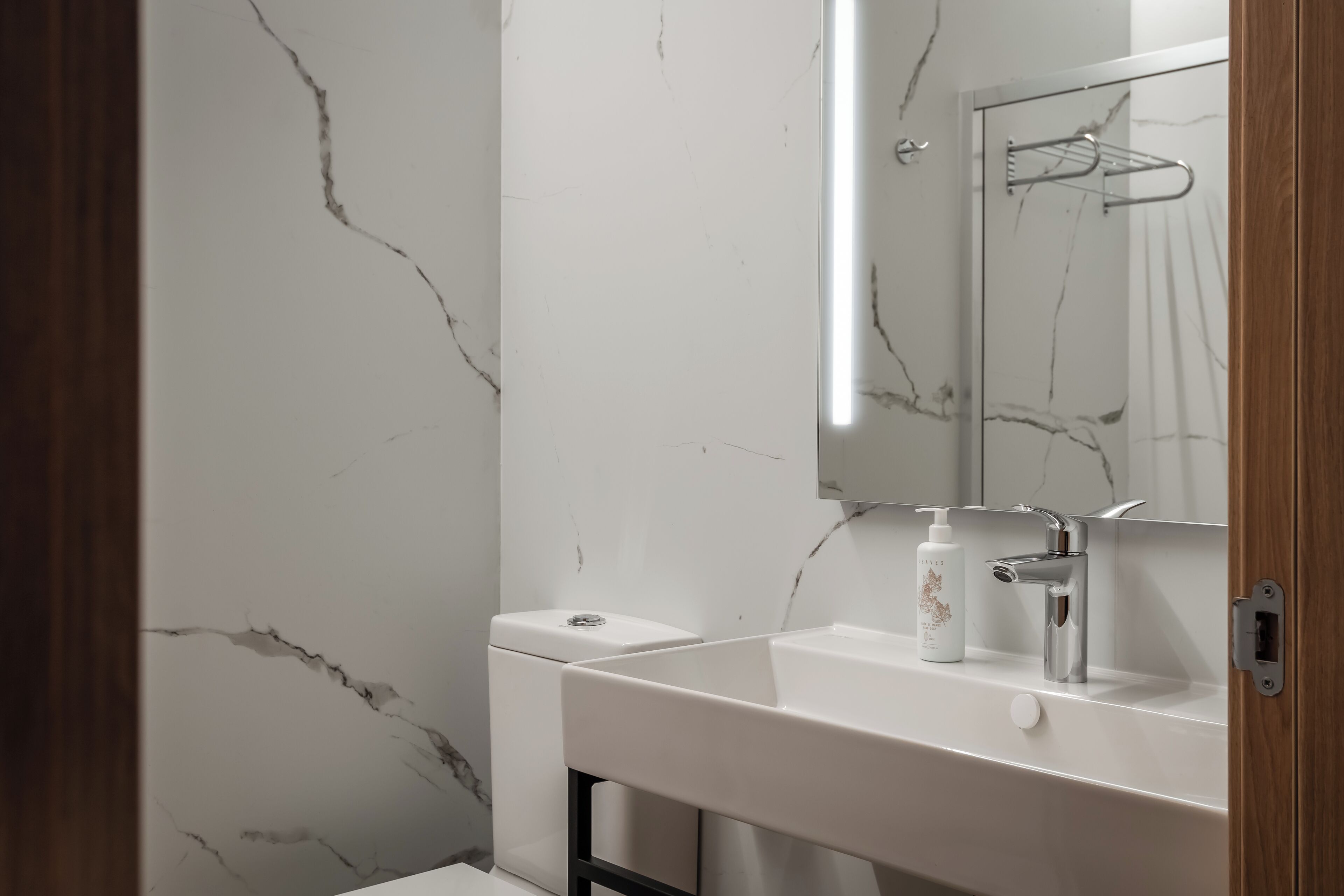 Un baño con pared de mármol blanco, un lavabo blanco con grifo cromado y un espejo sobre el lavabo.