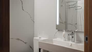Un baño con pared de mármol blanco, un lavabo blanco con grifo cromado y un espejo sobre el lavabo.