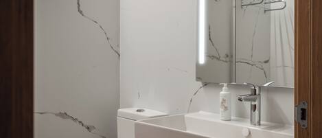 Un baño con pared de mármol blanco, un lavabo blanco con grifo cromado y un espejo sobre el lavabo.