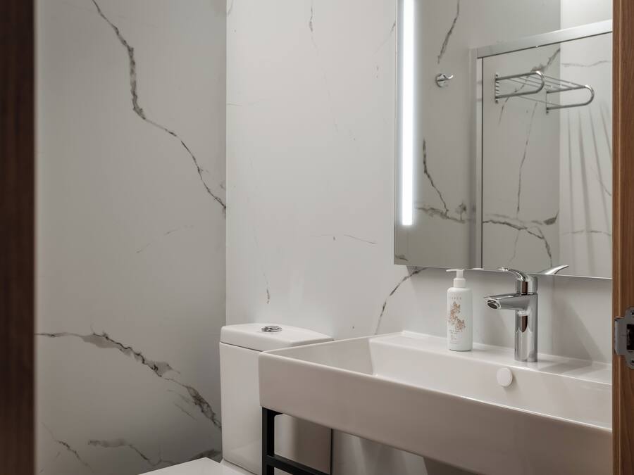 Un baño con pared de mármol blanco, un lavabo blanco con grifo cromado y un espejo sobre el lavabo.