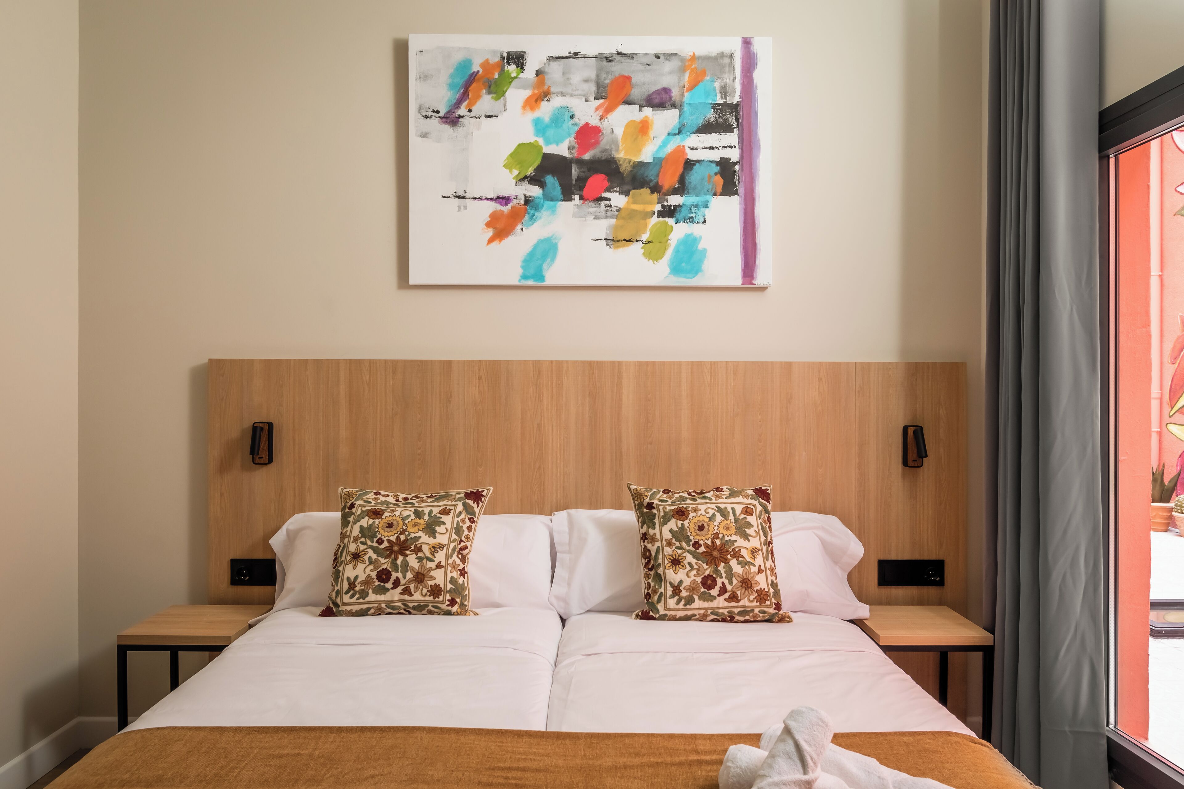 Habitación de hotel con cabecera de madera, dos almohadas estampadas con motivos florales, colcha blanca y un cuadro abstracto colorido en la pared.