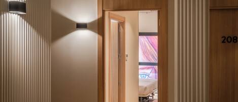 A hotel corridor with a wooden door leading to a room with a bed and a colorful artwork on the wall.