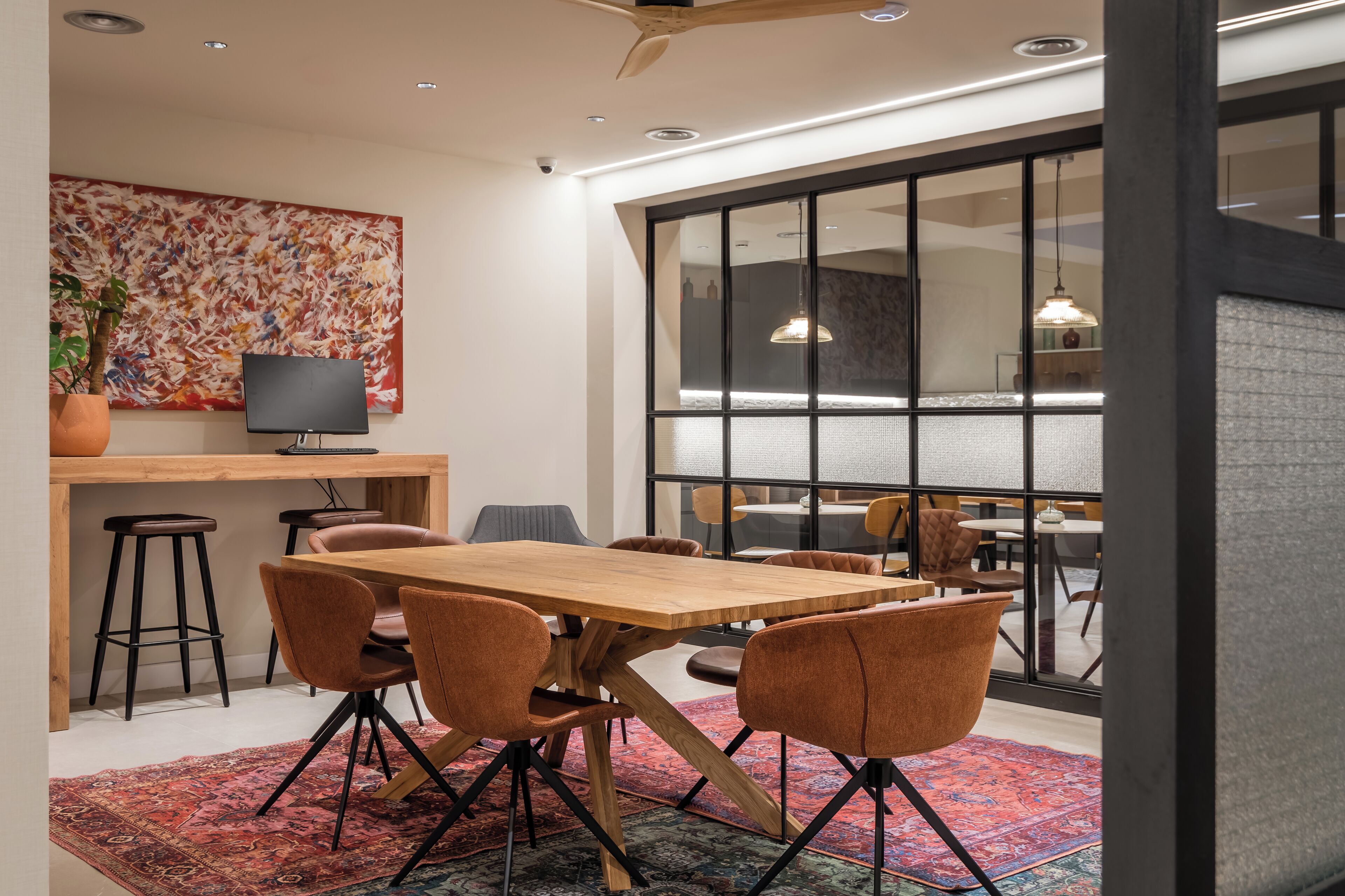 A modern dining area with a wooden table and brown chairs, a large abstract painting, and a view through a glass wall.