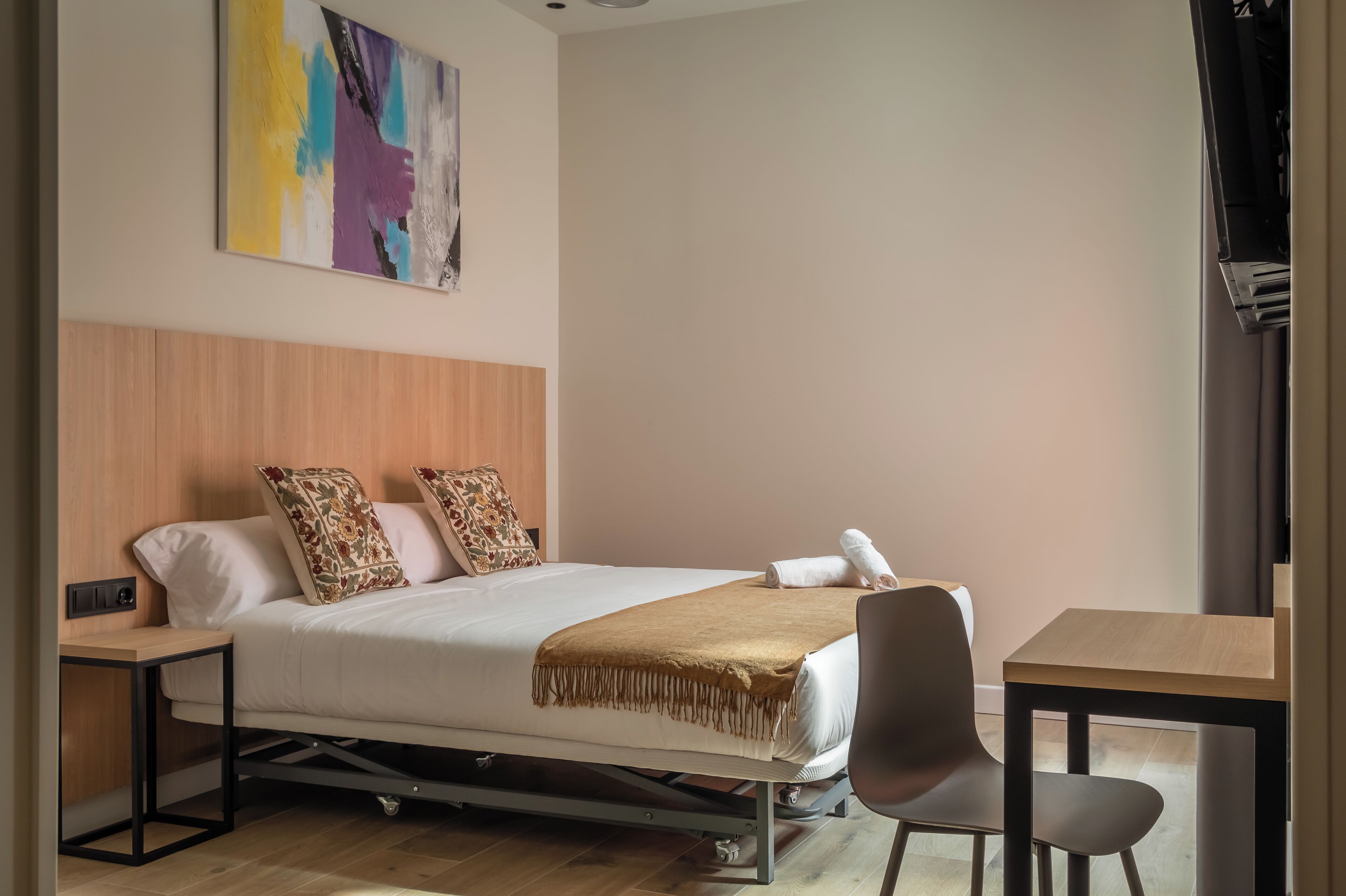 Una habitación de hotel ordenada con una cama, dos almohadas, una mesita de noche, una silla y una pequeña mesa.