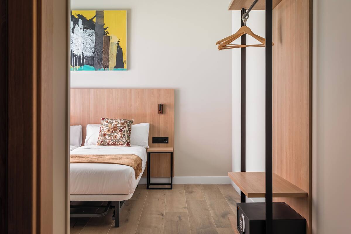 A modern hotel room with a wooden headboard, a bed with white bedding and a floral pillow, a bedside table with a lamp, and a wooden bench.