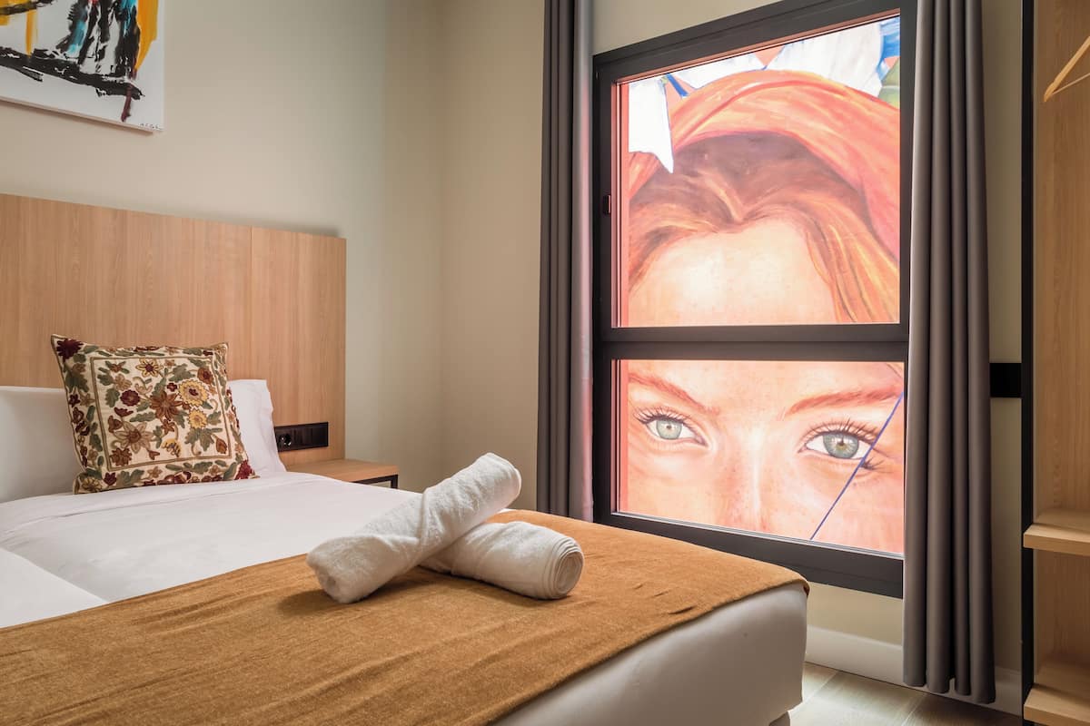 A hotel room with a bed, a window with a view, a wooden headboard, and a floral pillow.
