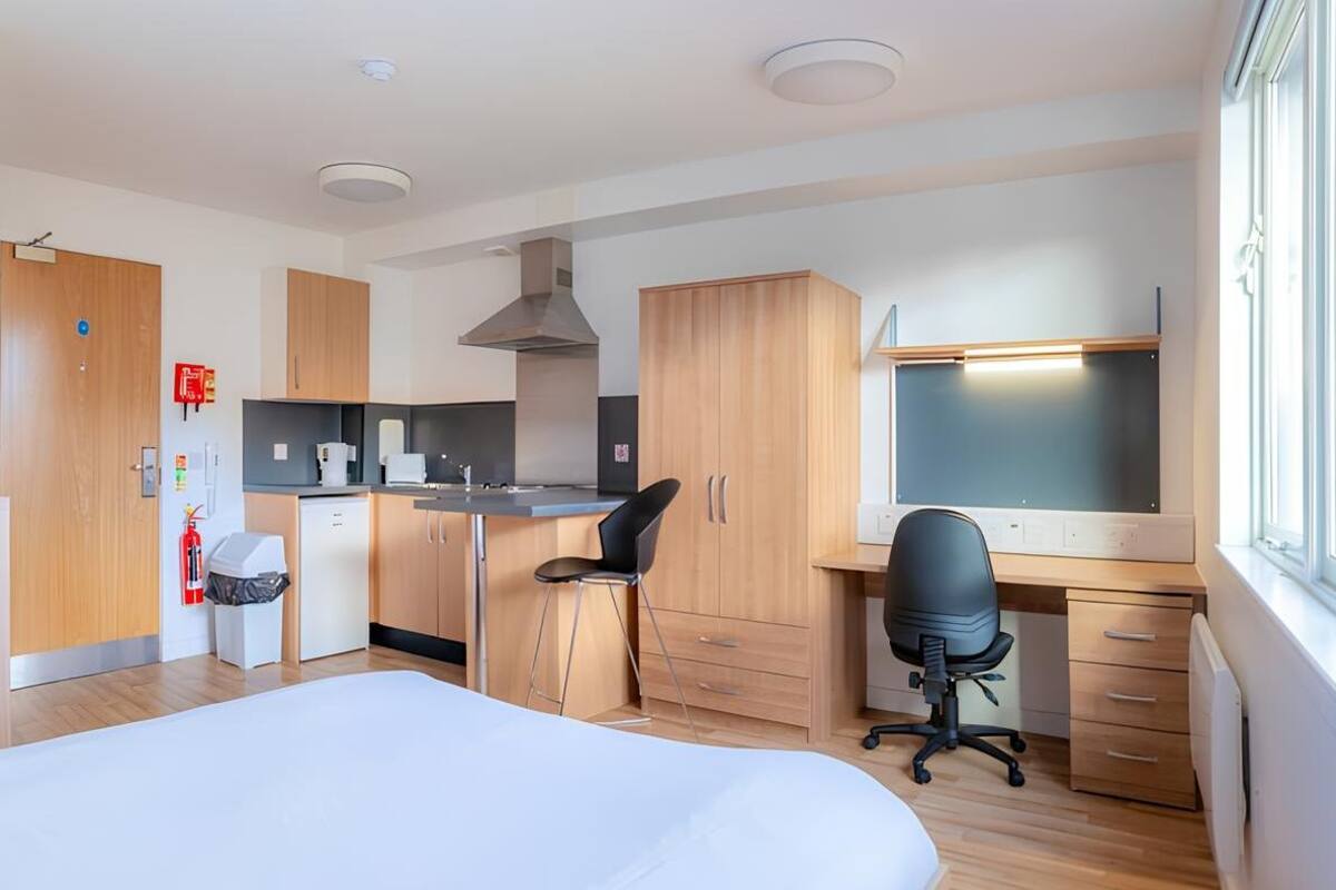 Ein kompaktes Hotelzimmer mit Bett, Küchenzeile, Schreibtisch mit Stuhl und Fenster.