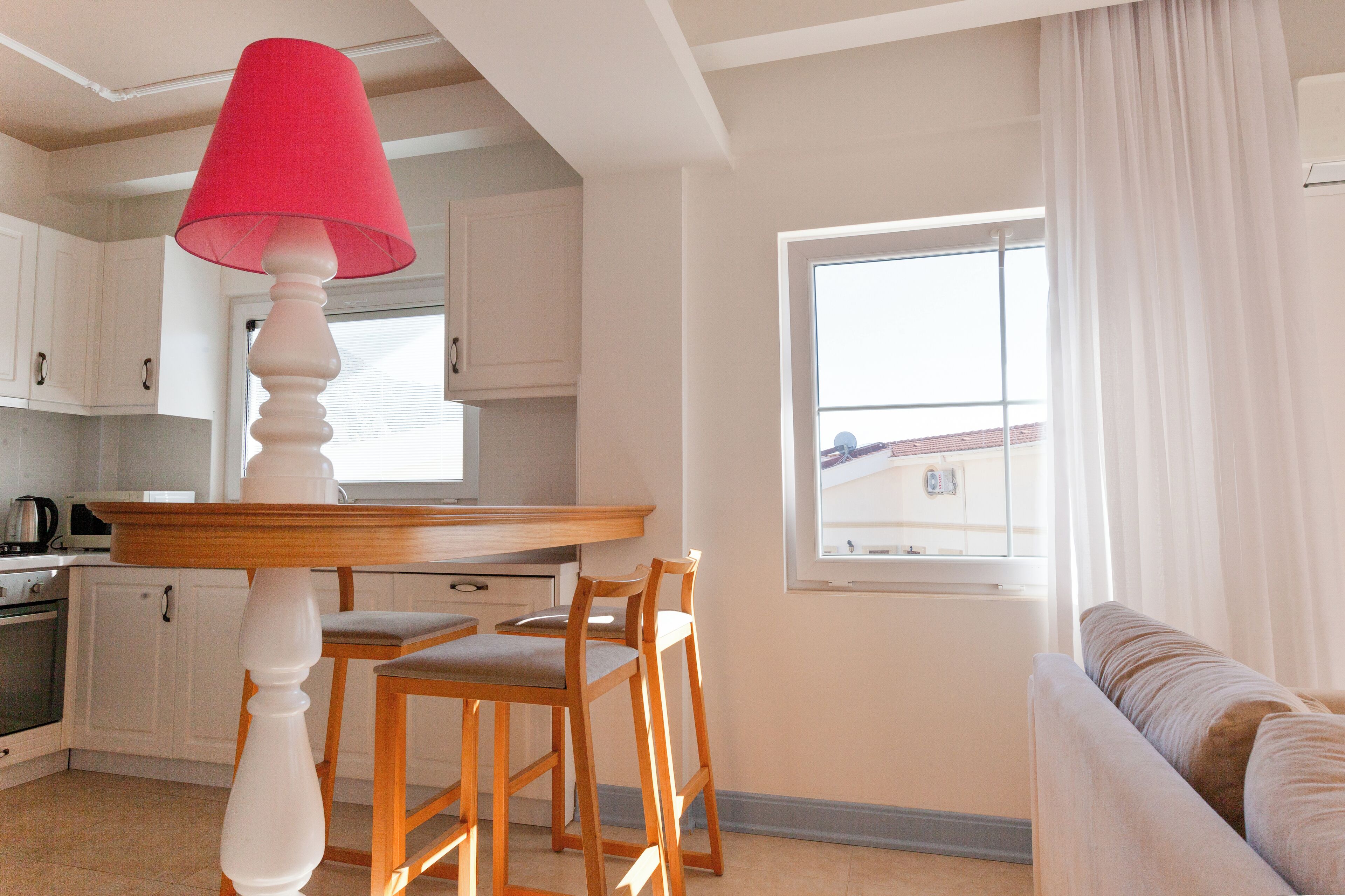 A kitchen with white cabinets, a wooden table, and a red lamp. There is a window with curtains and a view of a building outside.
