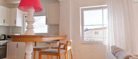 A kitchen with white cabinets, a wooden table, and a red lamp. There is a window with curtains and a view of a building outside.