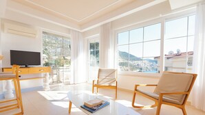 A modern living room with a flat-screen TV, a glass coffee table, and two rocking chairs. Large windows offer a view of a mountainous landscape. - Kalkan Suites Hotel (Kas)