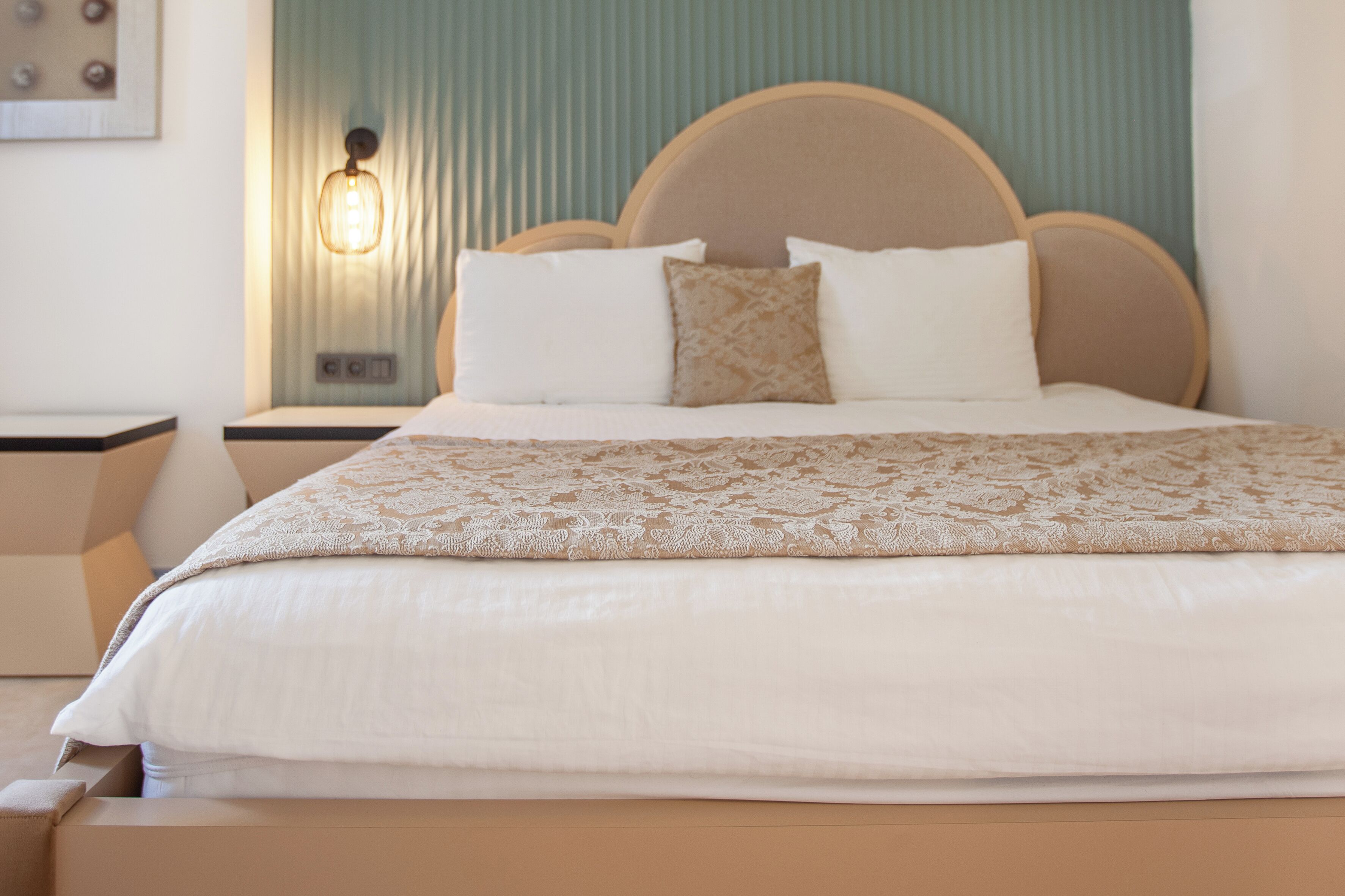 Una cama bien tendida con una colcha estampada y almohadas, un cabecero de madera y una lámpara fijada a la pared.