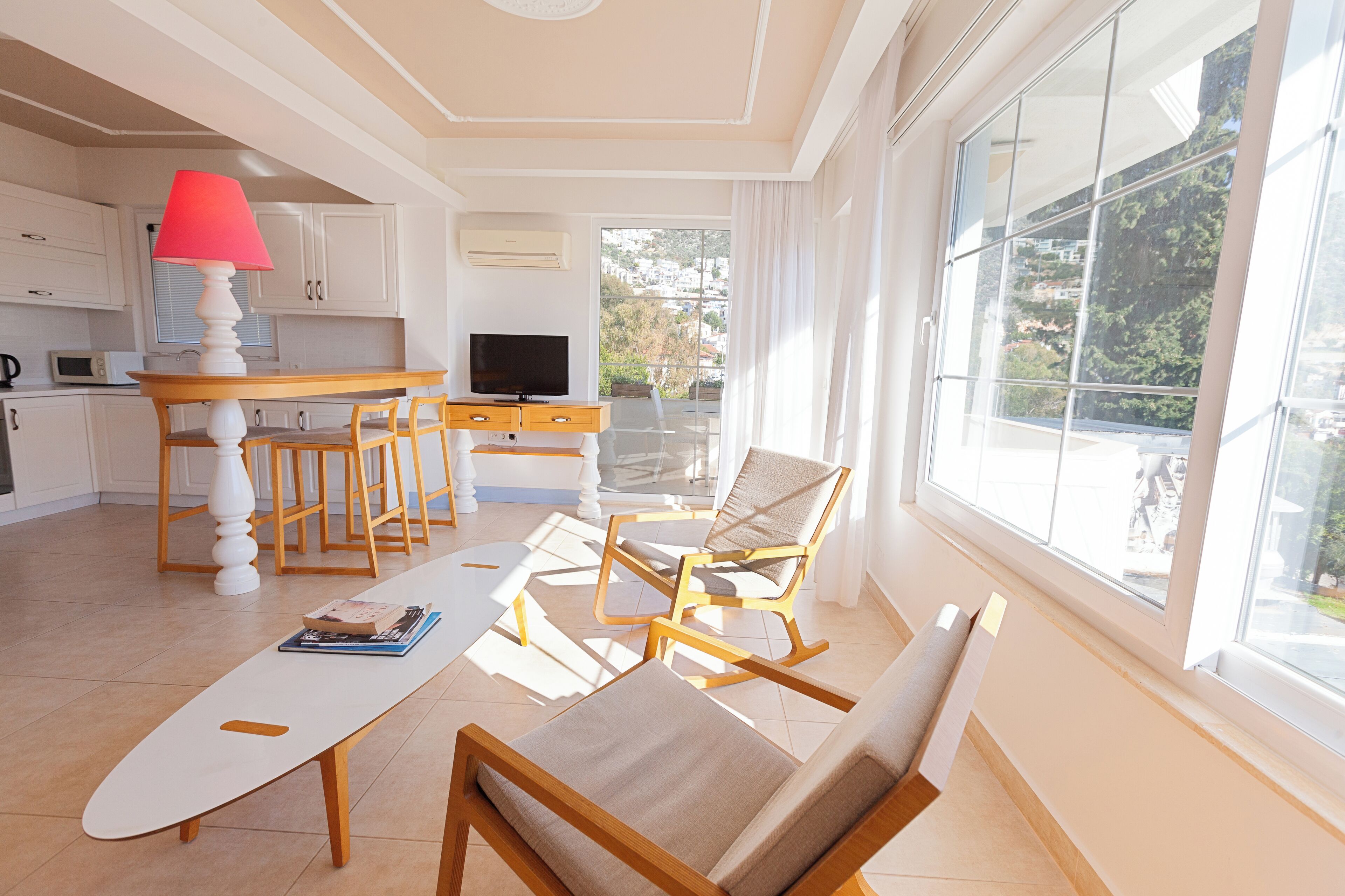 A modern living room with a sofa, a coffee table, and a TV. There is a kitchen area with white cabinets and a red lamp. The room has large windows with a view of greenery outside.