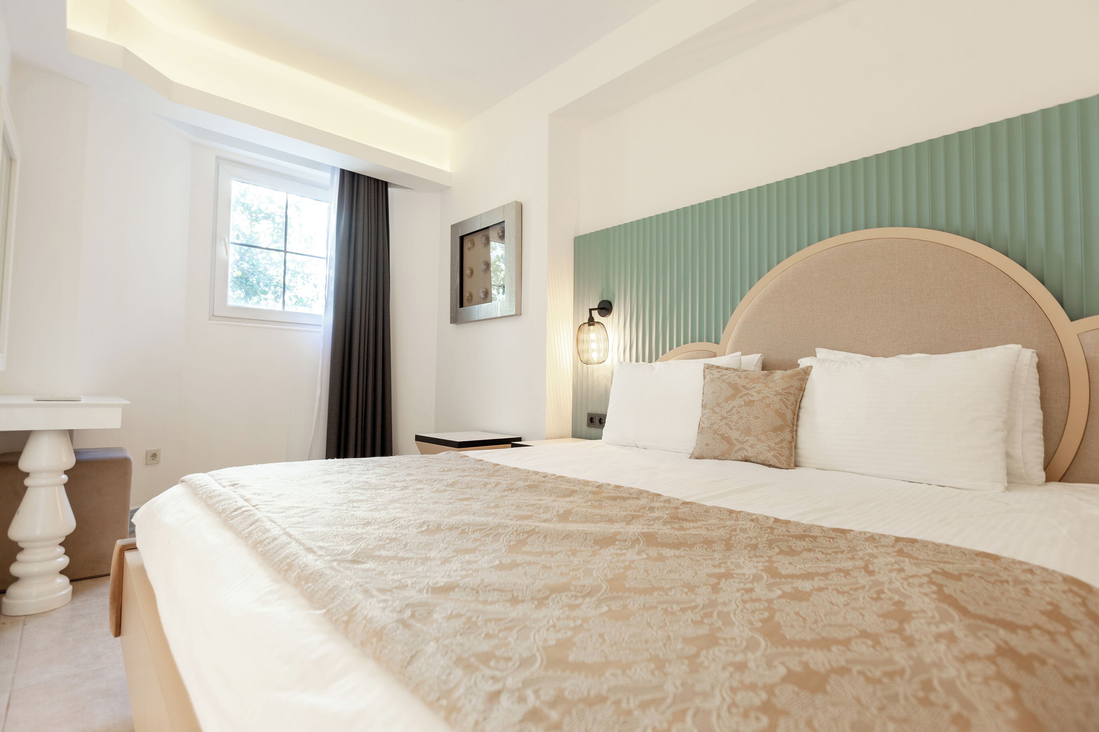 A hotel room with a large bed, a bedside table, a nightstand, a window with curtains, and a wall-mounted lamp.
