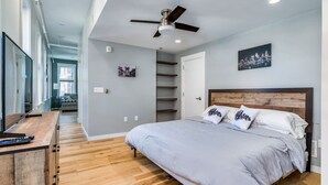 Een slaapkamer met een bed, een bureau, een plafondventilator en een stadsgezicht schilderij.