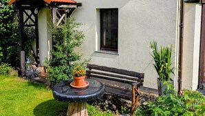 Een huis met een pannendak, een klein raam en een tuin met een bankje en potplanten.