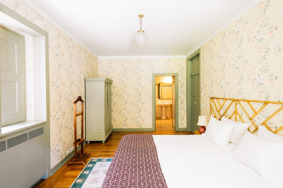Ein Schlafzimmer mit floraler Tapete, Holzboden, einem Bett mit weißen Leinentüchern, einem Nachttisch mit Lampe und einem Kleiderschrank.
