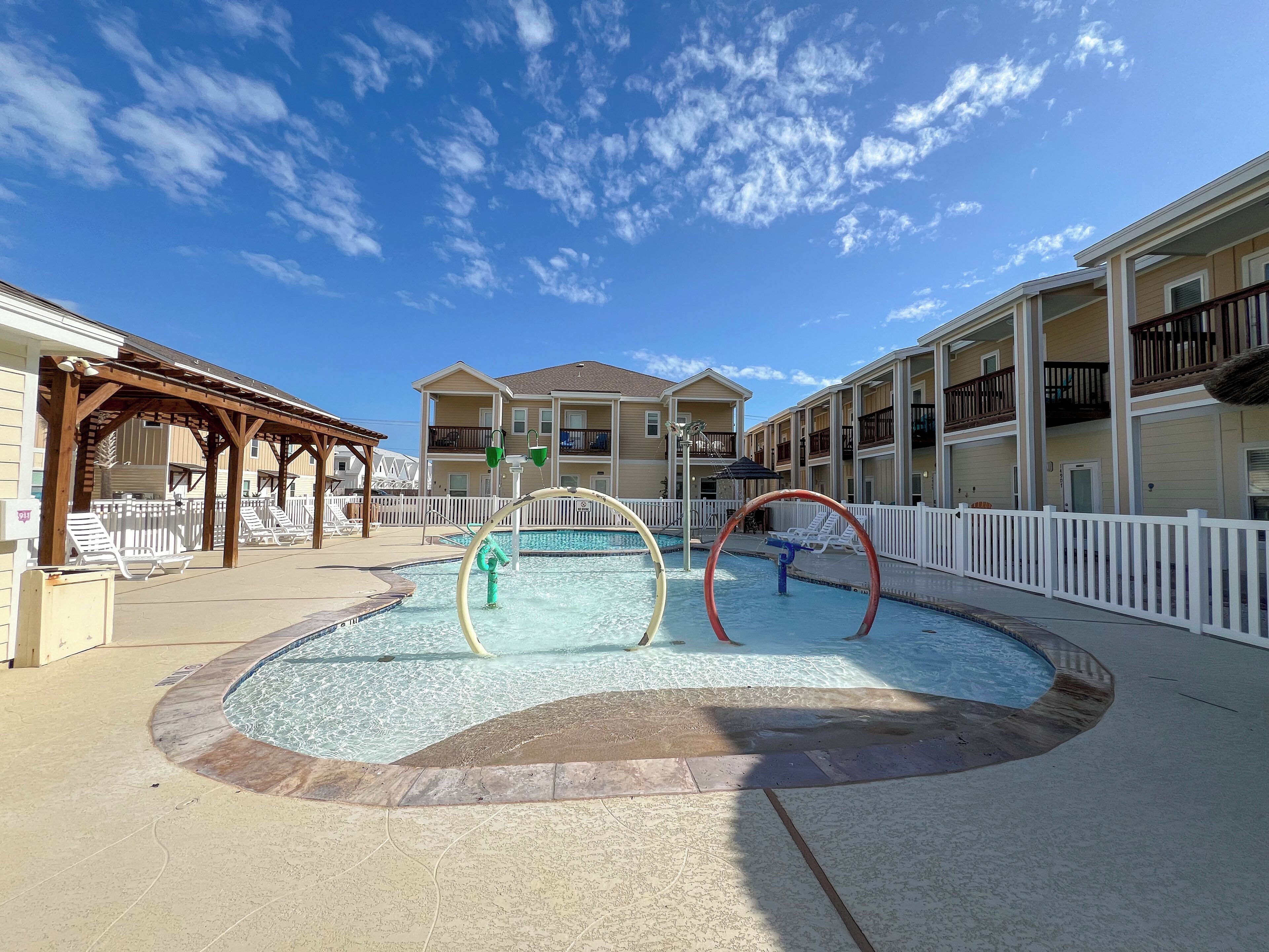 A pool area with a wooden deck, lounge chairs, and a building with balconies in the background.