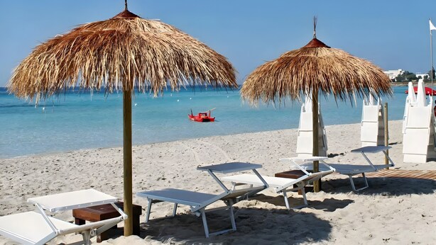 Une plage avec des parasols en chaume, des chaises longues blanches et un bateau rouge sur l’eau.