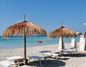 A beach with thatched umbrellas, white lounge chairs, and a red boat in the water.