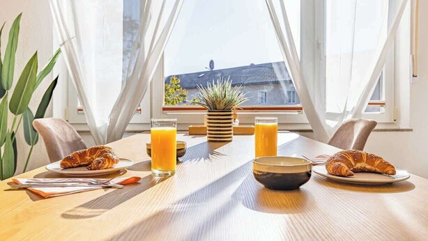 A breakfast setting with croissants, orange juice, and a bowl on a wooden table by a window with curtains. - SMS Homes I time out in Blaichach (Blaichach)