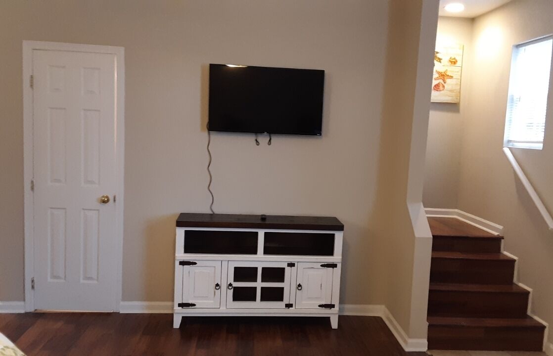 Ein Zimmer mit einer weißen Tür, einem an der Wand befestigten Flachbildfernseher, einer weißen Unterhaltungseinrichtung und einer Treppe mit Holzhäuschen.