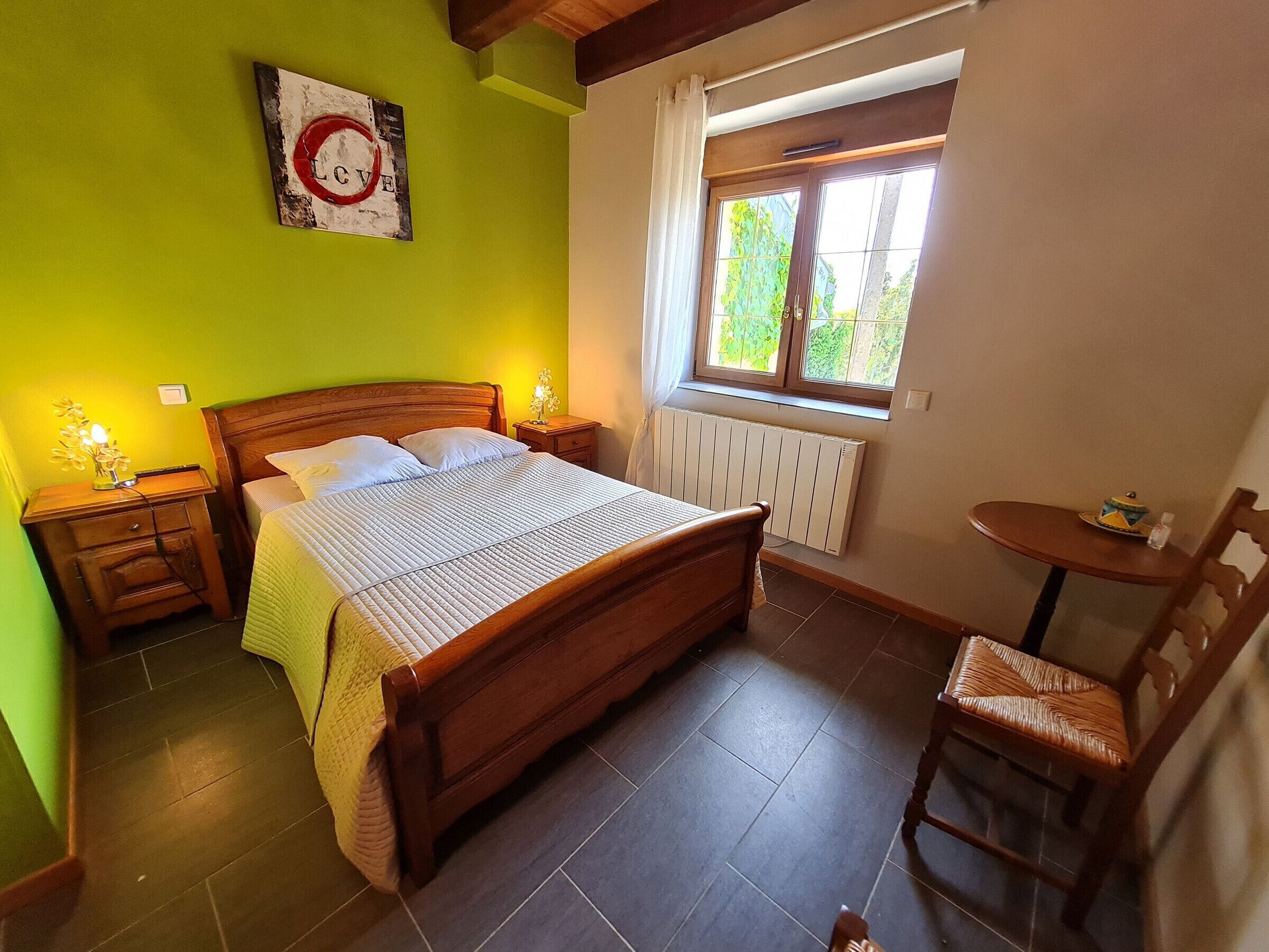 Een slaapkamer met een houten bed, een nachtkastje met een lamp, een stoel, een raam met uitzicht op groen en een radiator.
