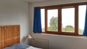 Une chambre à coucher avec une tête de lit en bois, un lit recouvert d’une couvre-lit à motifs, une fenêtre avec des rideaux bleus et un radiateur.