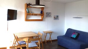 Un salon avec une table en bois, des chaises, un canapé bleu, un téléviseur fixé au mur, une étagère avec des livres et des objets décoratifs, et un luminaire suspendu.