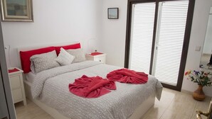 Ein Schlafzimmer mit einem Bett, Nachttischen, einem Fenster mit Jalousien und einer Vase mit Blumen.