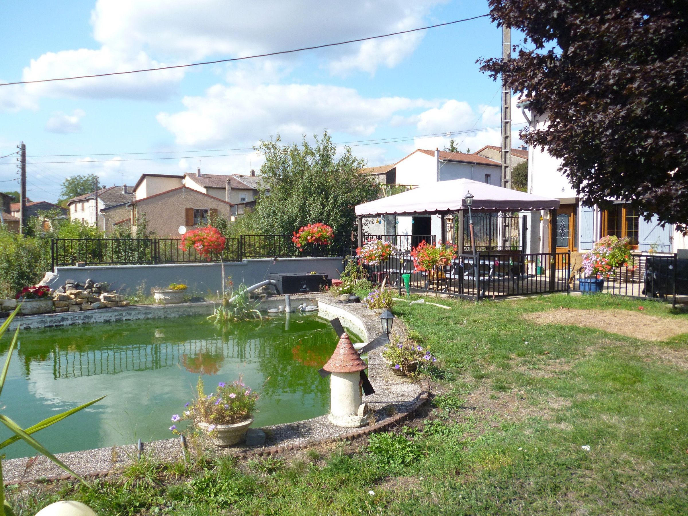 Un petit étang avec un pont, entouré d’un jardin où poussent différentes plantes et fleurs. On aperçoit des maisons au loin, et une clôture délimite la zone.