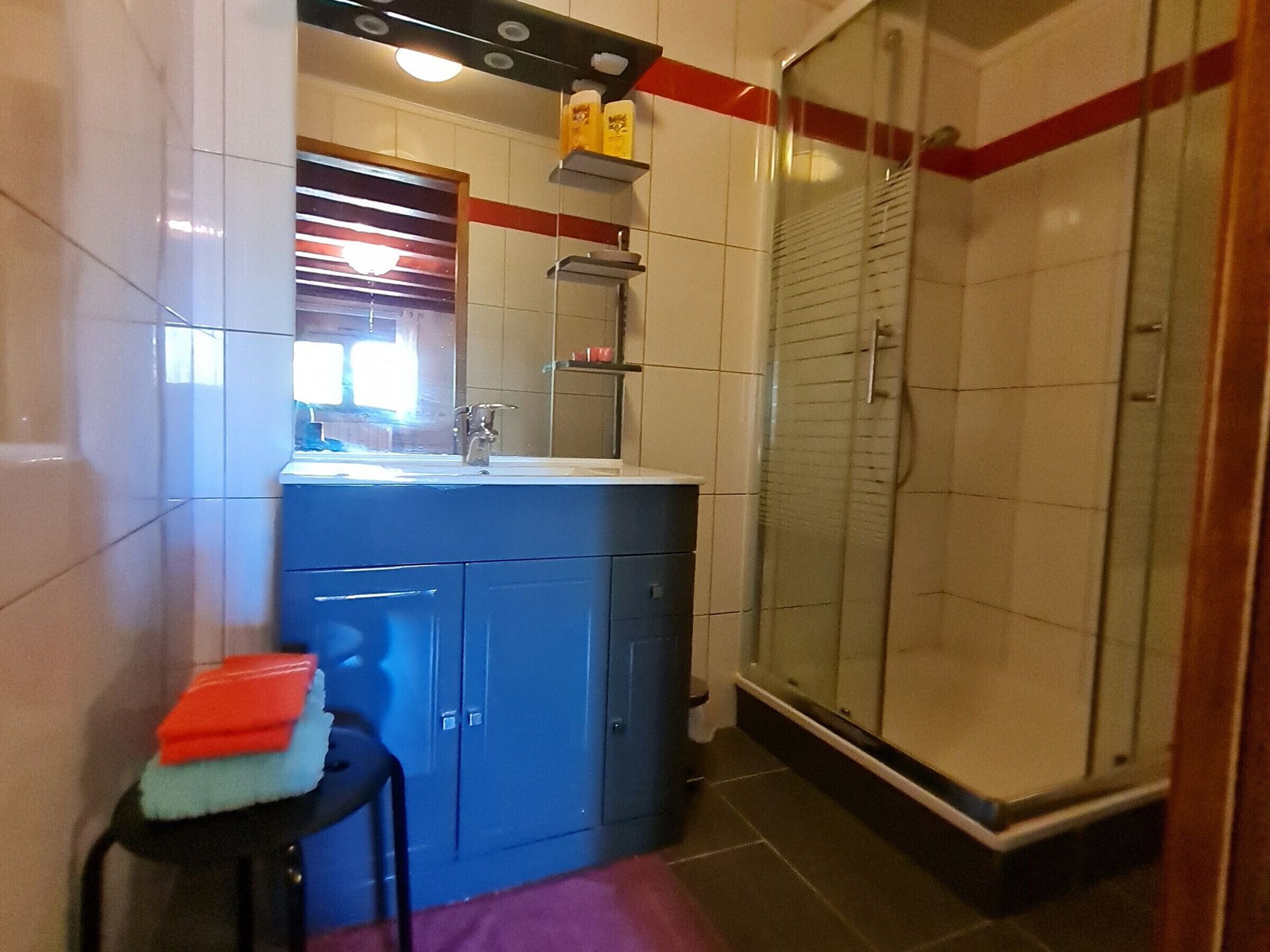 Een badkamer met een wastafel, spiegel en douche. Er staan toiletartikelen op het aanrecht en een handdoek op de vloer.