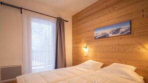 A bedroom with a wooden headboard, a bed with white bedding, a window with curtains, and a wall-mounted light fixture. - Valmorel: Apt for 6, 2 bdrm, parking, wifi, ski-in/ski-out (Valmorel)