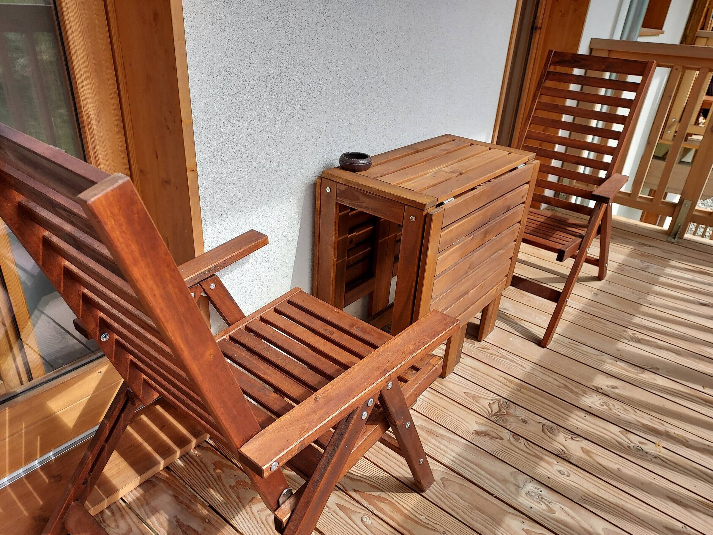 A wooden deck with a table and chairs, a window, and a white wall.
