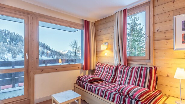 A cozy room with a wooden interior, a striped sofa, a small table, and large windows offering a view of a snowy landscape.