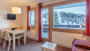A room with a wooden table and chairs, a small white table, a television mounted on the wall, and large windows with a view of a snowy landscape.