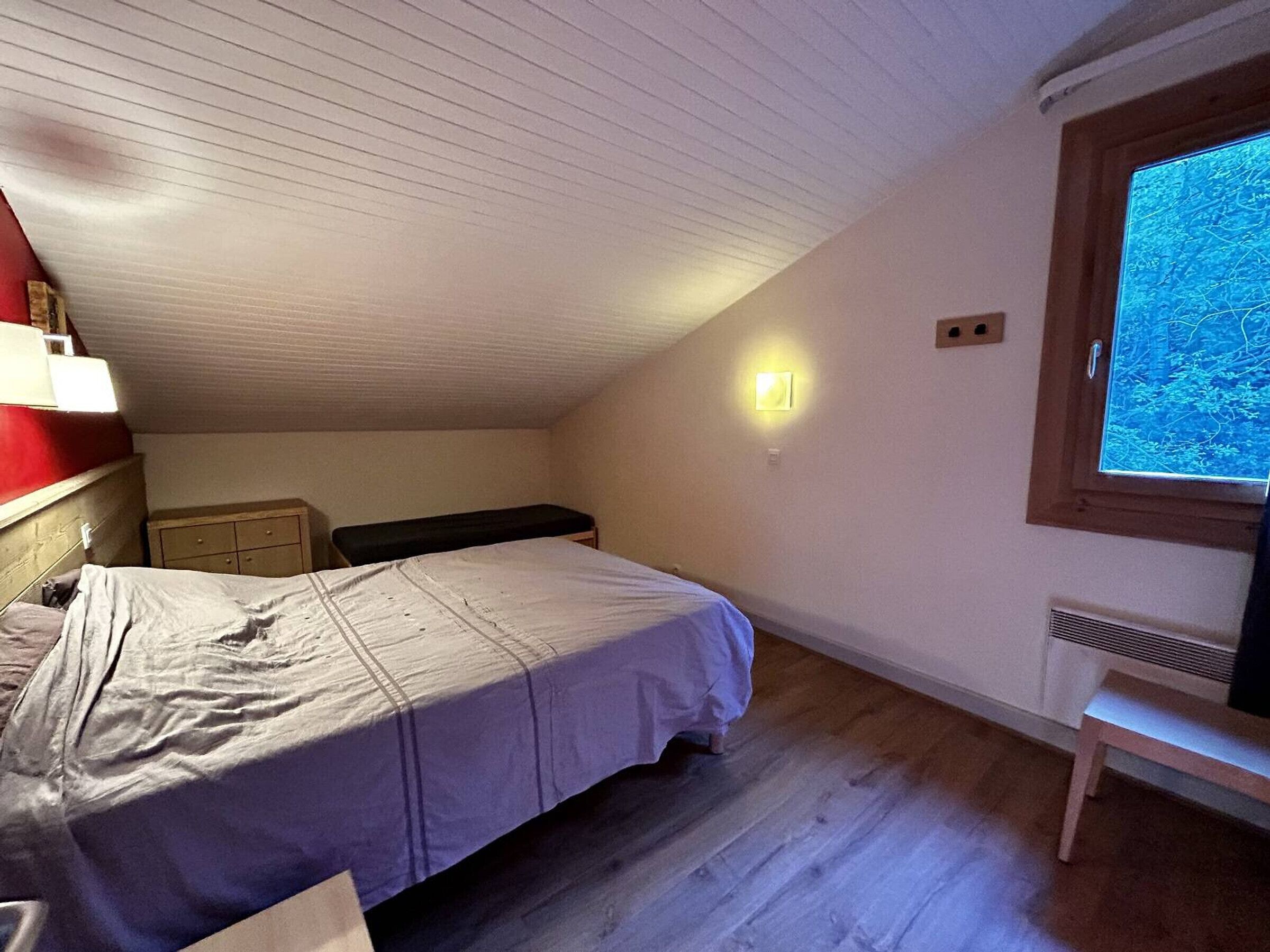 Et lille værelse med skråvæg, seng med hvid madras, et natbord, en bænk, en vægmonteret lampe og et vindue med udsigt til træer.