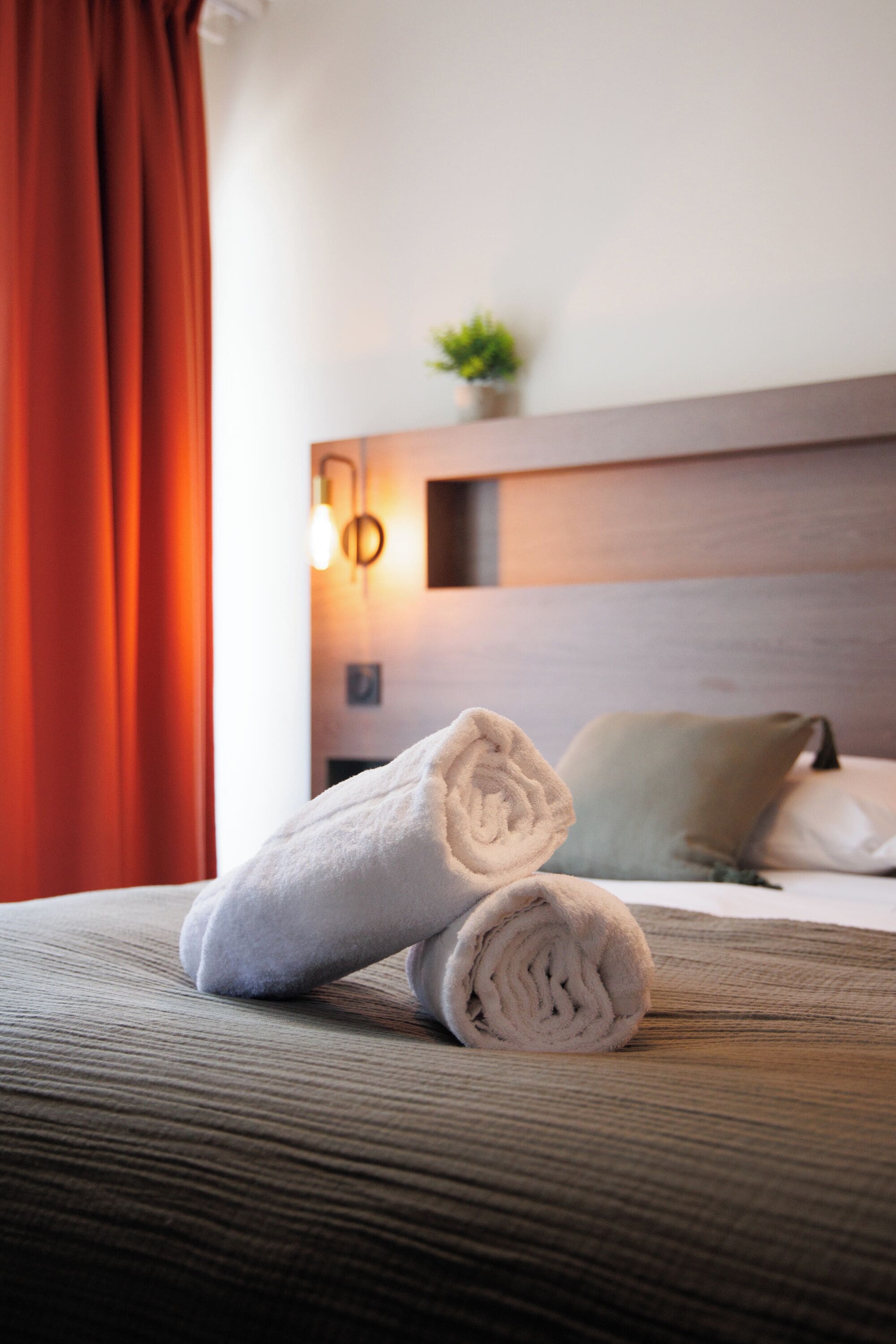 A neatly made bed with rolled towels, a wooden headboard, a small plant, and a wall-mounted light fixture.
