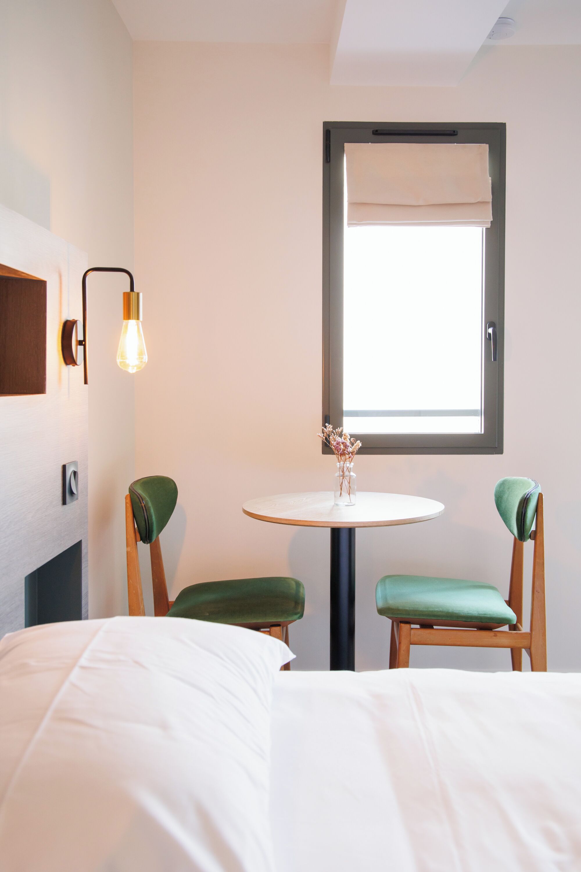 A hotel room with a bed, two chairs, a small table with a vase, a window with a blind, and a wall-mounted light fixture.