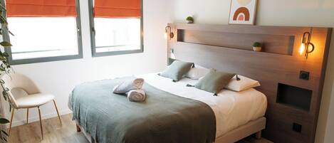 A modern bedroom with a bed, two windows with orange blinds, a bedside table with a lamp, and a small plant on the nightstand.