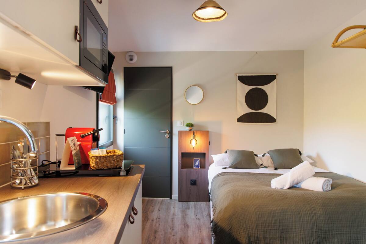 Une chambre d’hôtel compacte comprenant un coin cuisine, un lit avec des oreillers, une table de chevet et un miroir fixé au mur.