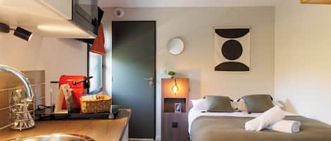 Una habitación de hotel compacta con kitchenette, una cama con almohadas, una mesita de noche y un espejo montado en la pared.