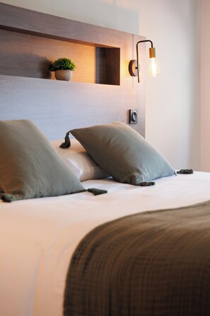 Una cama bien hecha con dos almohadas, una lámpara de pared y una pequeña planta en maceta.