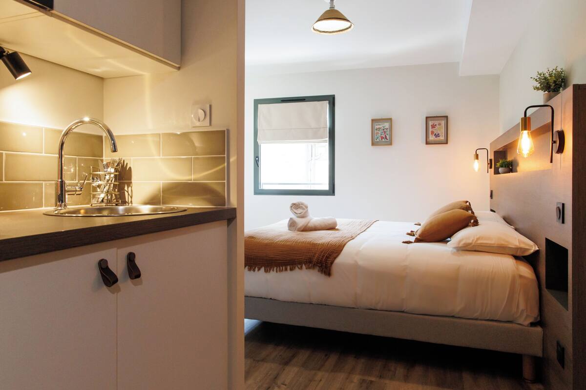 Une chambre d’hôtel moderne dotée d’une kitchenette, d’un lit recouvert d’un plaid marron, d’une fenêtre offrant une vue et d’un éclairage mural.