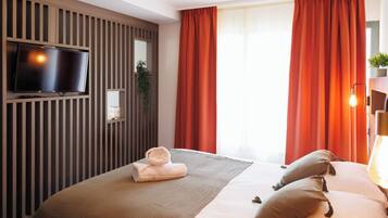 A modern hotel room with a flat-screen TV mounted on a wooden panel wall, a large bed with a beige blanket and pillows, red curtains, and a small potted plant on the windowsill.