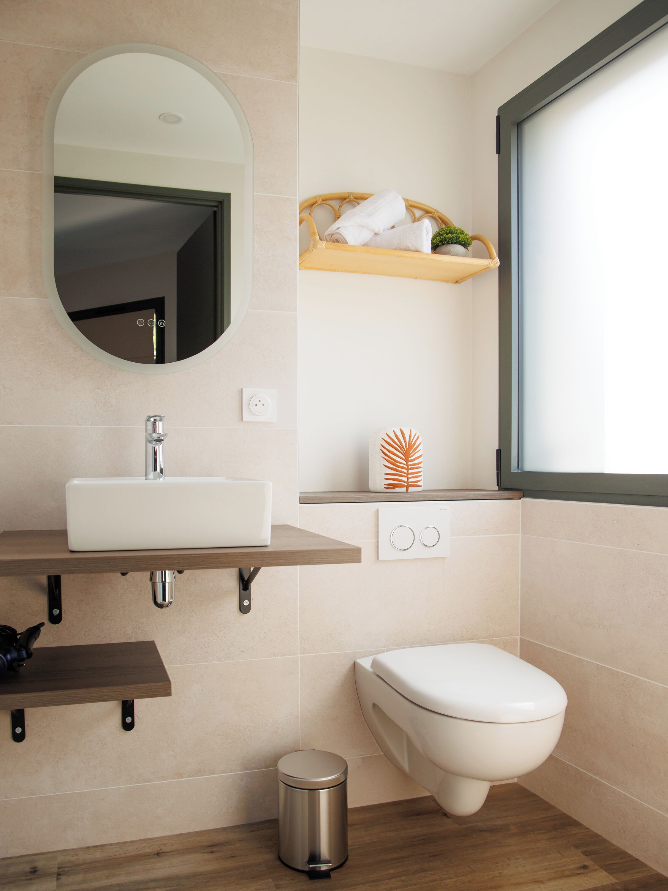 A modern bathroom with a white sink, mirror, and toilet. There is a shelf with towels and a decorative plant.