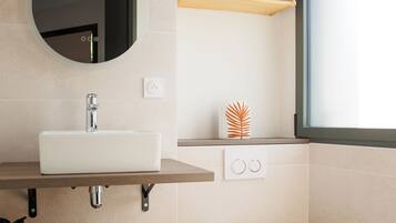 A modern bathroom with a white sink, mirror, and toilet. There is a shelf with towels and a decorative plant.